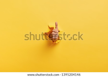 Image of unrecognizable man makes thumb up gesture, demonstrates approval or agreement, gestures through torn paper wall yellow background. Body language concept. Hand sign. Hole in wall. Like gesture