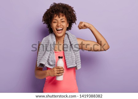 Horizontal shot of happy Afro woman shows biceps, satisfied after active training, holds bottle of water, enjoys healthy lifestyle, isolated over purple background. Sport and exercising concept