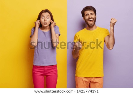Photo of surprised female plugs ears, shocked with noise, glad man clenches fists, celebrates triumph, yells loudly from happiness. Boyfriend and girlfriend stand over purple and yellow wall