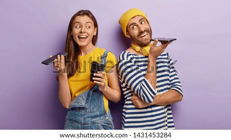 Indoor view of happy woman and man keep modern cell phones near mouthes, make voice call, enjoy nice conversation, drink hot beverage from disposable cup, stand back to each other over purple wall