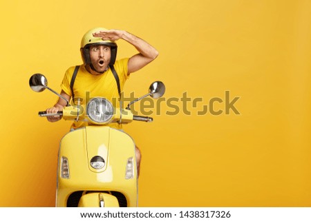 Stupefied male motorbike driver in ptotective helmet, keeps hand near forehead, focused with astonishment into distance, gazes something shocking, isolated on yellow wall. People and riding.