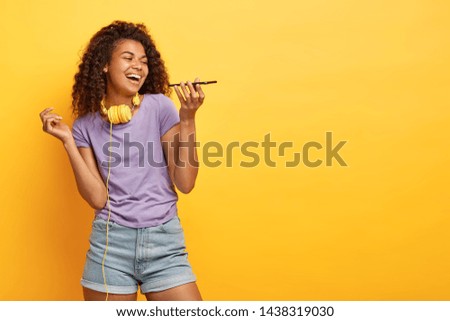 Studio shot of joyful Afro girl makes voice call, keeps modern cell phone near mouth, laughs gladfully, wears stylish clothing, has fun alone with gadget, poses over yellow studio wall, blank space