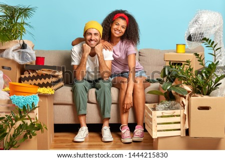 Optimistic multiethnic couple pose on sofa, settle in new apartment, being in high spirit, have much unpacked stuff at empty room with blue walls. Family, moving and starting new life concept