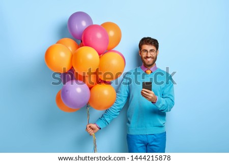 Glad man receives messages of congratulation, types feedback, holds modern smart phone, celebrates anniversary, holds big bunch of balloons, wears blue sweater and trousers, smiles at camera