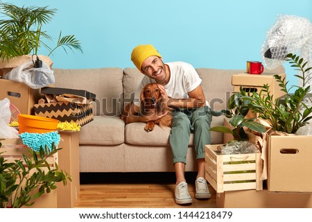 New beginning concept. Happy guy embraces favourite dog, brought boxes with things for new home, rests on sofa, owns apartment, lives together with domestic pet. Overjoyed man settles in abode