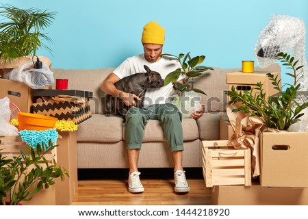 Fashionable man poses on cozy sofa with favourite pet, move together in new home, room in mess with boxes. Male homeowner spends free time with dog in bought abode. Starting new life concept