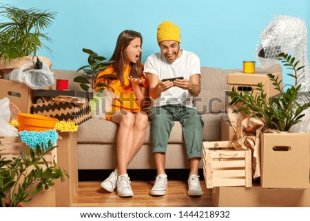 Discontent wife being angry with husband addicted to technologies, plays online games on smartphone, move in new dwelling, have much work, asks to help with unpacking belongings. Estate purchasing