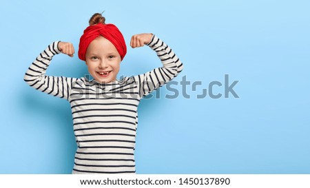 Strong happy small girl raises arms and shows biceps, has good physical shape, being active, wears red headband, black and white striped sweater, poses indoor. Positive preschool child feels powerful