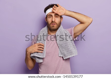 Weariness, sport concept. Tired man carries towel on shoulders, touches forehead, being sweaty after long cardio training, poses in gym over purple background. Motivated sportsman rests after workout