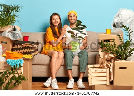 Delighted woman and man sit on sofa with household objects, buy luxury house to start new family life. Smiling husband holds pot of houseplant, mirthful wife leans at shoulder. First home concept