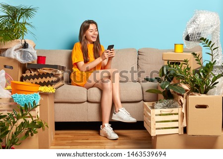 Moving in new house. Joyful pretty European woman rejoices buying expensive flat, sends text messages for friends via mobile phone, big boxes packages around, blue background, changes place of living