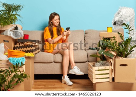 Moving concept. Cheerful young female model surrounded with lot of boxes, sits on comfortable sofa in room, happy this place is her, chats in social networks, searches for new design for house