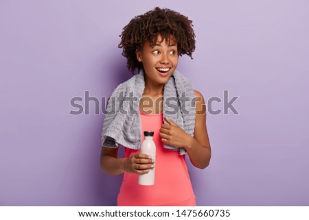 Joyful glad woman looks happily aside, poses with towel on shoulders, drinks fresh water after exhausting exercising, takes break, has dehydration of body. Athletic Afro girl rests after jogging
