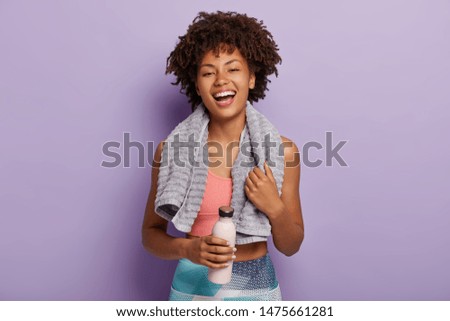 Smiling fitness woman in top and leggings takes break after training, holds bottle of water, wipes sweat with towel, being energetic runner or jogger, feels thirsty. People, wellness, vitality concept