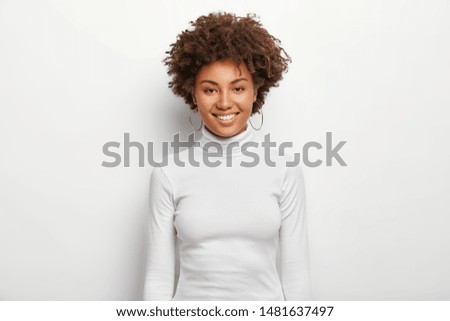 Photo of charismatic lovely woman with curly hair, has fun, toothy smile on face, satisfied after successful deal, looks at camera, wears white casual clothes, poses in studio. Happy emotions