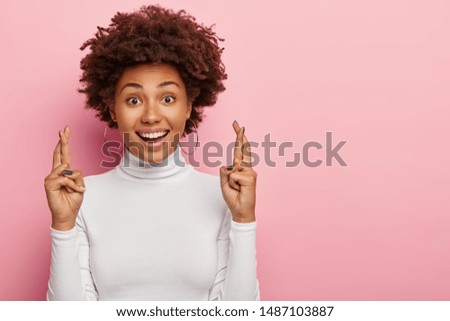 Headshot of glad smiling curly woman crosses fingers with positive expression, believes in good luck and success, looks hopefully straightly at camera, isolated over pink wall with blank space aside