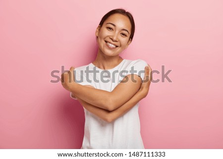 Love yourself. Pretty glad Asian girl embraces herself, feels comfort and care, tilts head, wears white t shirt, has no make up, isolated over rosy wall, thinks about lover, wants be in his warm arms