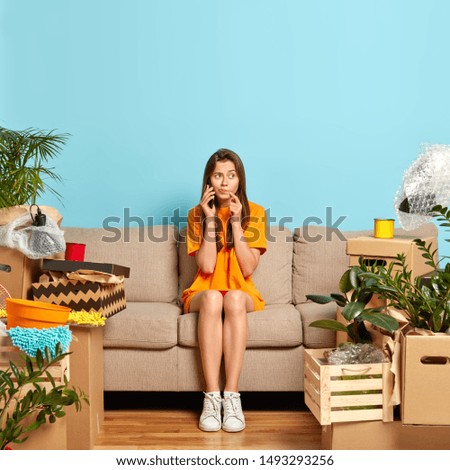 Photo of serious woman calls someone via smartphone, sits on comfortable sofa, shares news about buying new apartment, surrounded with personal belongings, relaxes in new home. Moving concept