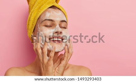 Feminity and hygiene concept. Young delighted female washes face with soap, smiles happily, closes eyes from satisfaction, cleans skin, poses against pink background with blank space for your promo