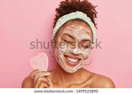 Beautiful optimistic Afro American woman cleanses face with foam, refreshes skin, has well cared complexion, holds heart shaped sponge for beauty procedures, stands bare shoulders with closed eyes