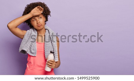 Sporty girl with Afro hair wipes forehead, being sweaty, dressed in casual vest, holds bottle with fresh water, has regular training for keeping fit, wears towel on shoulders, isolated on purple wall