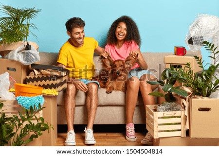 Starting new life in recently bought apartment. Happy diverse woman and man have fun with dog, play with its ears, pose on sofa, have to bring everything in order, enjoy first day at new home