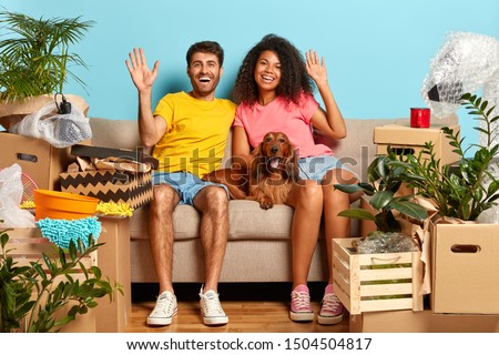 Indoor shot of happy diverse family couple wave at camera, sit on comfortable sofa, pedigree dog lies near, celebrate moving day, have many boxes with belongings to unpack, being in good mood