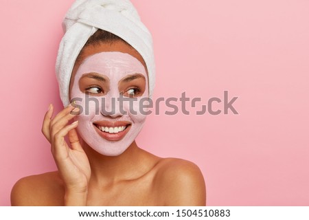Close up shot of lovely African American woman has delicate sooth skin, wears cream mask on face to reduce acnes, has healthy complexion, hygienic treatments wears white wrapped towel on head