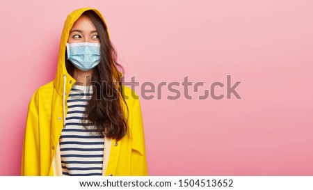 Photo of brunette young ill woman wears protective medical mask, has infectional disease, dressed in yellow raincoat, covers head with hood, looks aside, isolated on pink background, copy space area