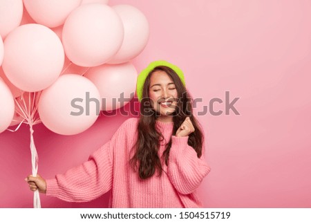 Photo of happy pleased brunette Asian lady stands with balloons, enjoys cool party with friends, wears beret and loose jumper, celebrates anniversary, poses against pink background. Festive event