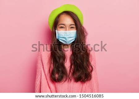 Lovely dark haired Asian lady has epidemic disease, wears protective medical mask, green beret and sweater, poses against pink background, has infection, stands indoor. Health care and people concept