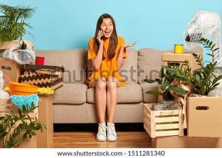Positive young girl discusses something actively, gesture and holds mobile phone near ear, wears loose orange t shirt and sneakers, shares impressions about moving in new dwelling. Changing home