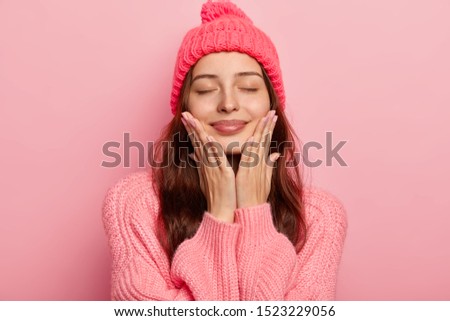 Portrait of pleased European woman touches both cheeks with palms, wears knitted hat and sweater, closes eyes with satisfaction, imagines future date with boyfriend enjoys winter time isolated on pink