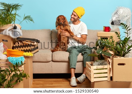 Happy man pets pedigree dog, pose on sofa, move to new house, lot of packing cartons around, rejoice buying modern flat, rest after moving in. Homeowner with pet spend first day at new dwelling