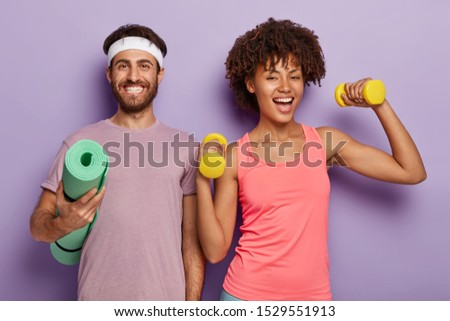 Improve your muscles. Sporty woman trains with dumbbells, has cheerful look, her husband stands near, holds rolled up fitness mat, enjoy athletic training together, isolated on purple background
