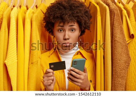 Sad frustrated Afro woman makes payment via online wallet, holds credit card and modern cellphone, unhappy to have little money on her account, stands near different yellow clothes hanging on hangers