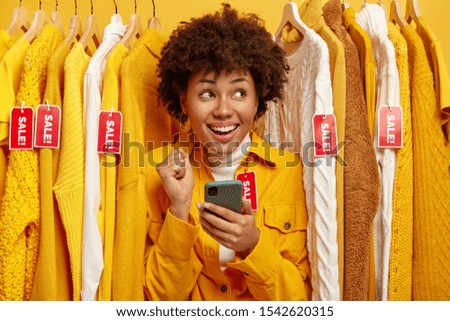 Sale, fashion, discounts and online shopping concept. Overjoyed dark skinned woman chooses clothes in clothing store, rejoices big sales, holds mobile phone for making payment via special app