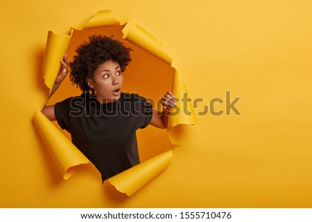 Photo of surprised Afro woman in black t shirt gasps from amazement, looks with scared face expression aside, dressed in black t shirt, unexpected to see something horrible, poses in torn paper wall