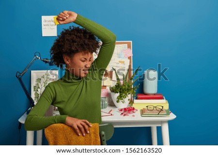Displeased curly woman smells unpleasant stinch under armpit, wears green turtleneck, sits over coworking space, prepares for examination session in own cabinet, feels tired. Copy space area.