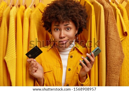Displeased unaware woman with curly hairstyle, unable to pay all sum of money for clothes, holds plastic card and modern mobile phone, poses against plain yellow jumpers on hangers. Paying online