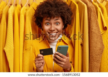 Positive mixed race shopaholic stands near rack with clothes, uses credit card and smartphone for making purchasing, smiles broadly as get big sale for being regular customer of fashion store
