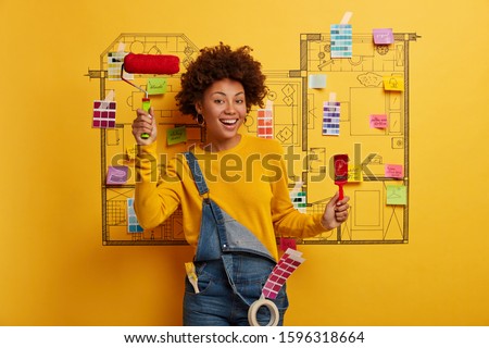 Renovation at home. Positive African American woman holds paint brush and roller, busy with painting, thinks about house improvement, dressed in jean overalls, creative sketch on yellow wall