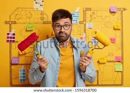 Doubtful bearded painter man paints walls with roller, chooses between red and yellow color, busy with renovation and refurbishment, stands against house sketch, sticky notes, yellow background