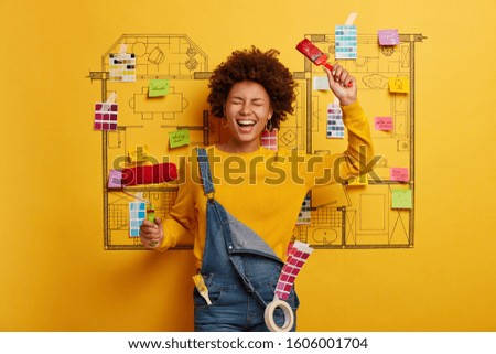 Joyous optimistic Afro American woman rejoices finishing painting, holds paintbrush and roller, enjoys renovation work, wears denim dungarees, remodels house in modern style, improves dwelling