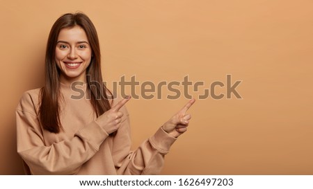 Look, advertise here! Dark haired pretty woman with appealing smile gives positive attitude towards good deal, expresses her recommendation while pointing on right side, wears brown sweater.