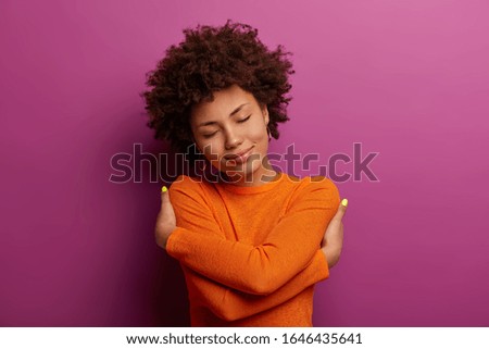 Lovely pretty woman embraces herself, feels good, comfortable and fullfilled, has high self esteem, tilts head and closes eyes, being egoisitc person, wears orange jumper isolated on purple background 商業照片 © 