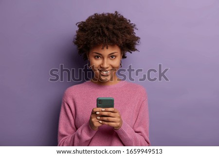 Portrait of lovely ethnic woman holds modern mobile phone, uses electronic device on surfing web, looks positively at camera, connected to wireless internet, wears casual sweater, poses indoor