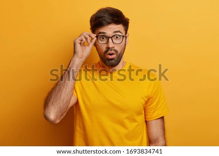 Man in stupor learns bad shocking news, keeps hand on rim of spectacles, has eyes popped out, cannot believe in fake relevation, dressed casually, isolated on yellow background, feels astounded Foto d'archivio © 
