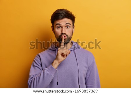 Mysterious bearded young man makes shush gesture, asks to keep secret safe, says keep voice down, quiet please, touches finger over lips, dressed in purple hoodie, isolated on yellow background