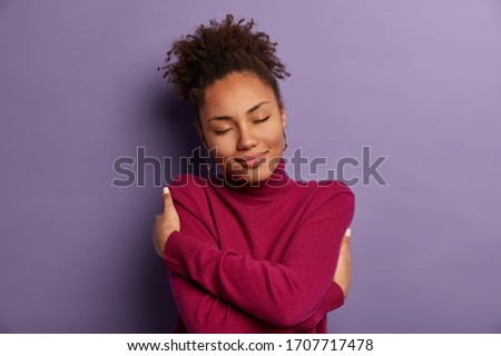 I love myself. Tender romantic woman embraces own body, hugs herself, closes eyes from pleasure, wears soft turtleneck just for cold weather, feels comfort, stands indoor against purple wall